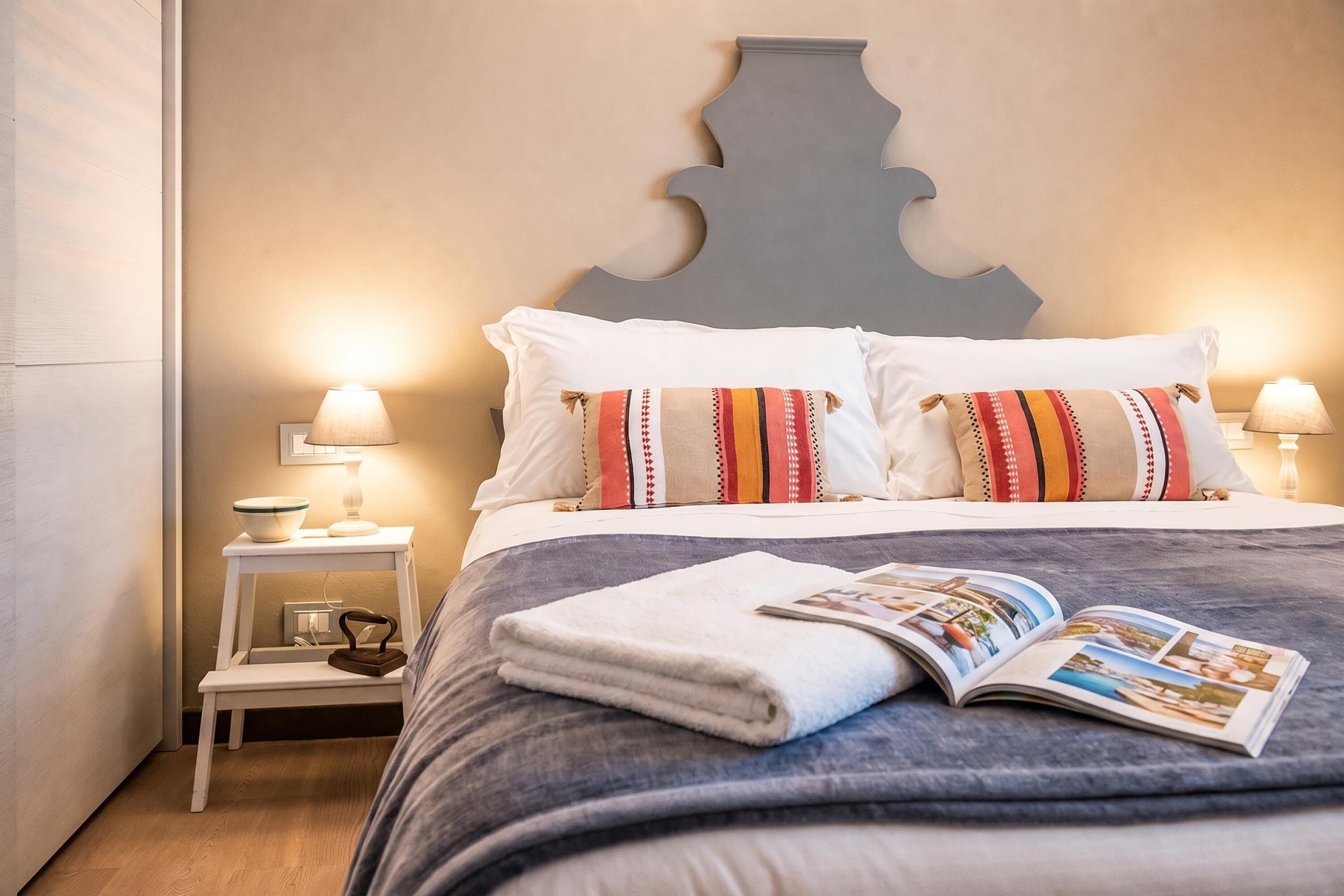 Un lit bien fait, avec un magazine à portée de main, une petite table de chevet avec une lampe et une tête de lit décorative.