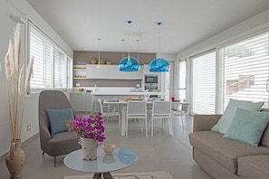 A modern living room with a sofa, armchair, and coffee table. There is a dining area with a table and chairs. The kitchen is visible in the background with white cabinets and a microwave.