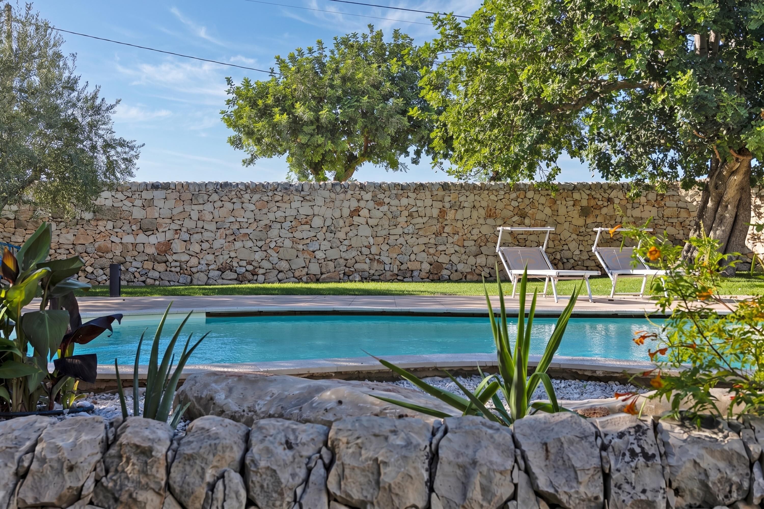 A swimming pool with a stone wall, two lounge chairs, and surrounding greenery.