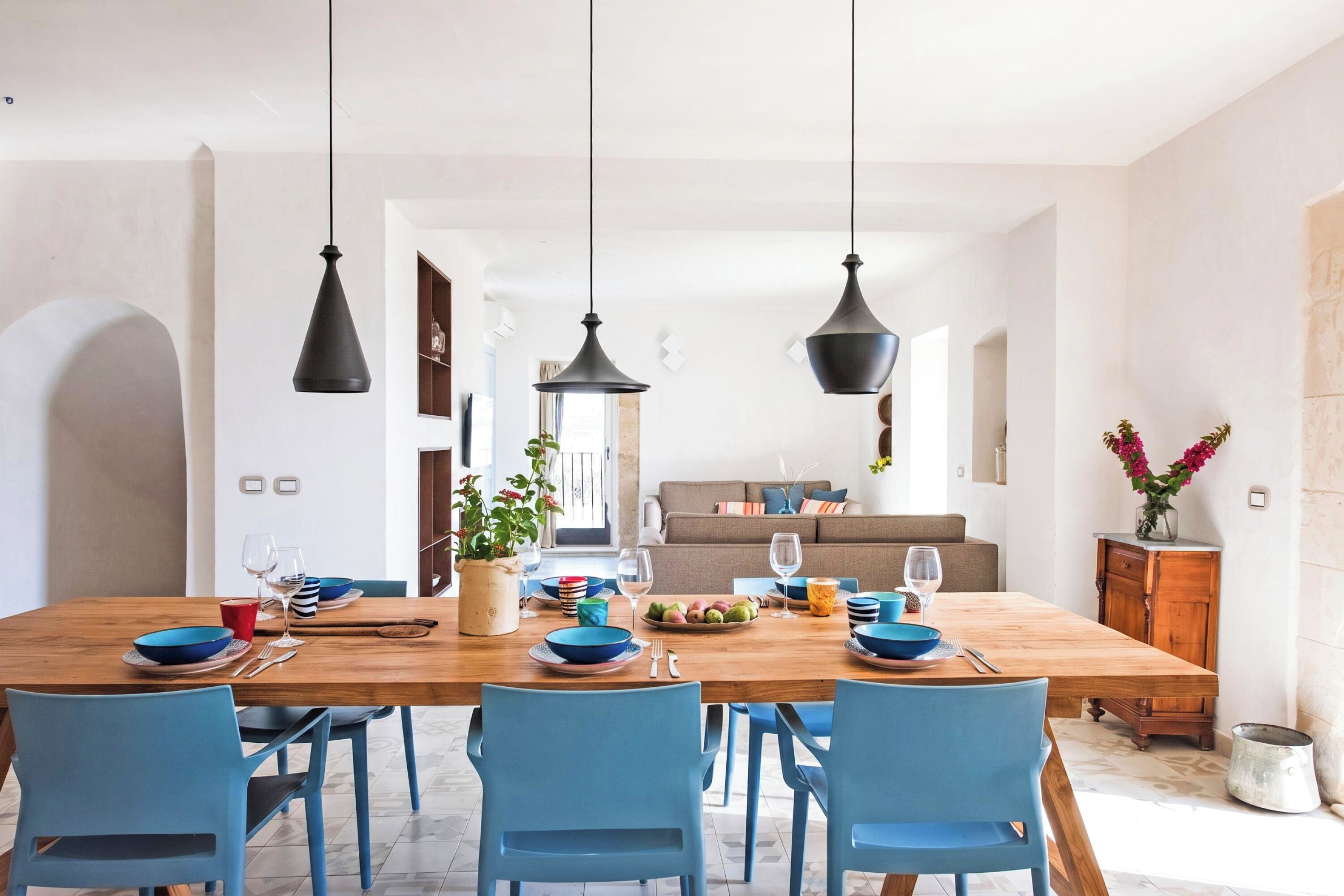 A dining area with a wooden table, blue chairs, black pendant lights, and a view into a living room with a sofa and a plant.