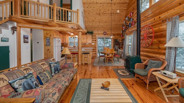 A log cabin interior with a loft bedroom, a dining area, and a living room furnished with a sofa and chairs. - Log Cabin Hideaway- Cozy Mountain Getaway (Arnold)