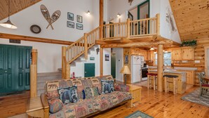 A spacious living area with a wooden staircase, a sofa with patterned cushions, and a kitchen area with wooden cabinets. - Log Cabin Hideaway- Cozy Mountain Getaway (Arnold)