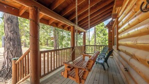 A wooden deck with a swing chair and a table, surrounded by a wooden railing and a large tree. - Log Cabin Hideaway- Cozy Mountain Getaway (Arnold)