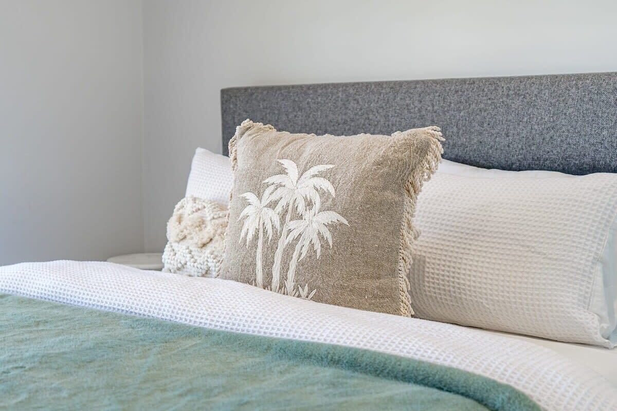 A bed with a textured white cover, a beige pillow with palm tree embroidery, and a grey headboard.