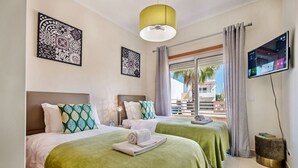 A hotel room with two beds, green bedding, a flat-screen TV, artwork on the walls, and a balcony with curtains.