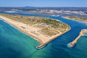 An aerial view of a coastal area with a beach, a marina, and a peninsula with buildings and vegetation.