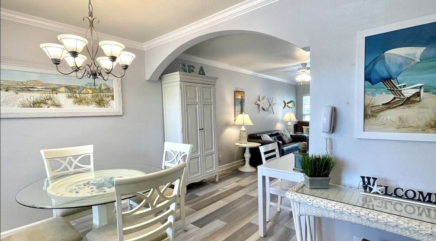 A dining area with a glass table, white chairs, a chandelier, and a 'Welcome' sign.