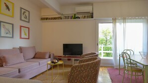 Ein Wohnzimmer mit einer Couch, einem Fernseher, einem gemusterten Sessel, einem Esstisch mit Stühlen und einem Fenster mit Blick auf Grünfläche.