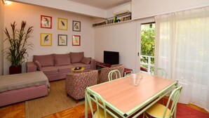 Ein Wohnzimmer mit Sofa, Couchtisch, Esstisch mit Stühlen, Fernseher und Bücherregal.