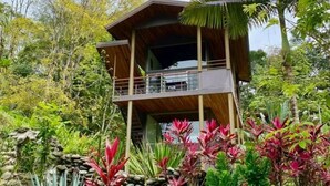 A two-story wooden cabin with a balcony, surrounded by lush tropical vegetation. - A Sloth Riverfront Villa with Jacuzzi Pool (Bahía Ballena)