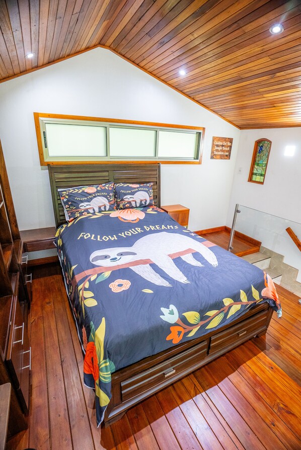 Un dormitorio con techo inclinado, suelo de madera, cama con colcha de diseño alegre con perezosos, mesita de noche y escalera con barandilla de vidrio.