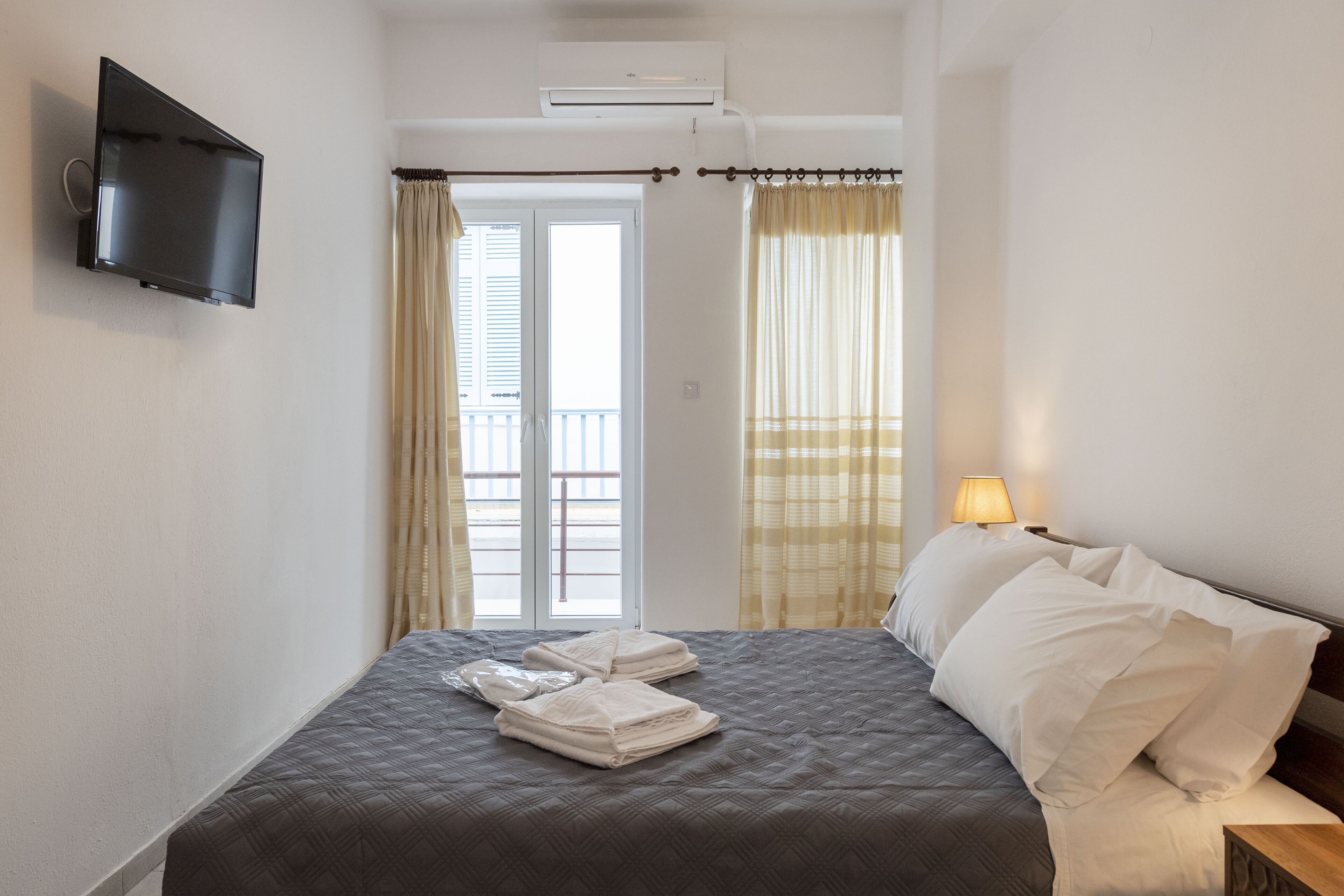 A hotel room with a bed, a TV mounted on the wall, a window with curtains, and a bedside lamp.