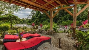 A covered outdoor seating area with red cushions, surrounded by a garden with rocks, plants, and a view of a river. - Toucan Riverfront Villa with Pool Near A Beach (Uvita)