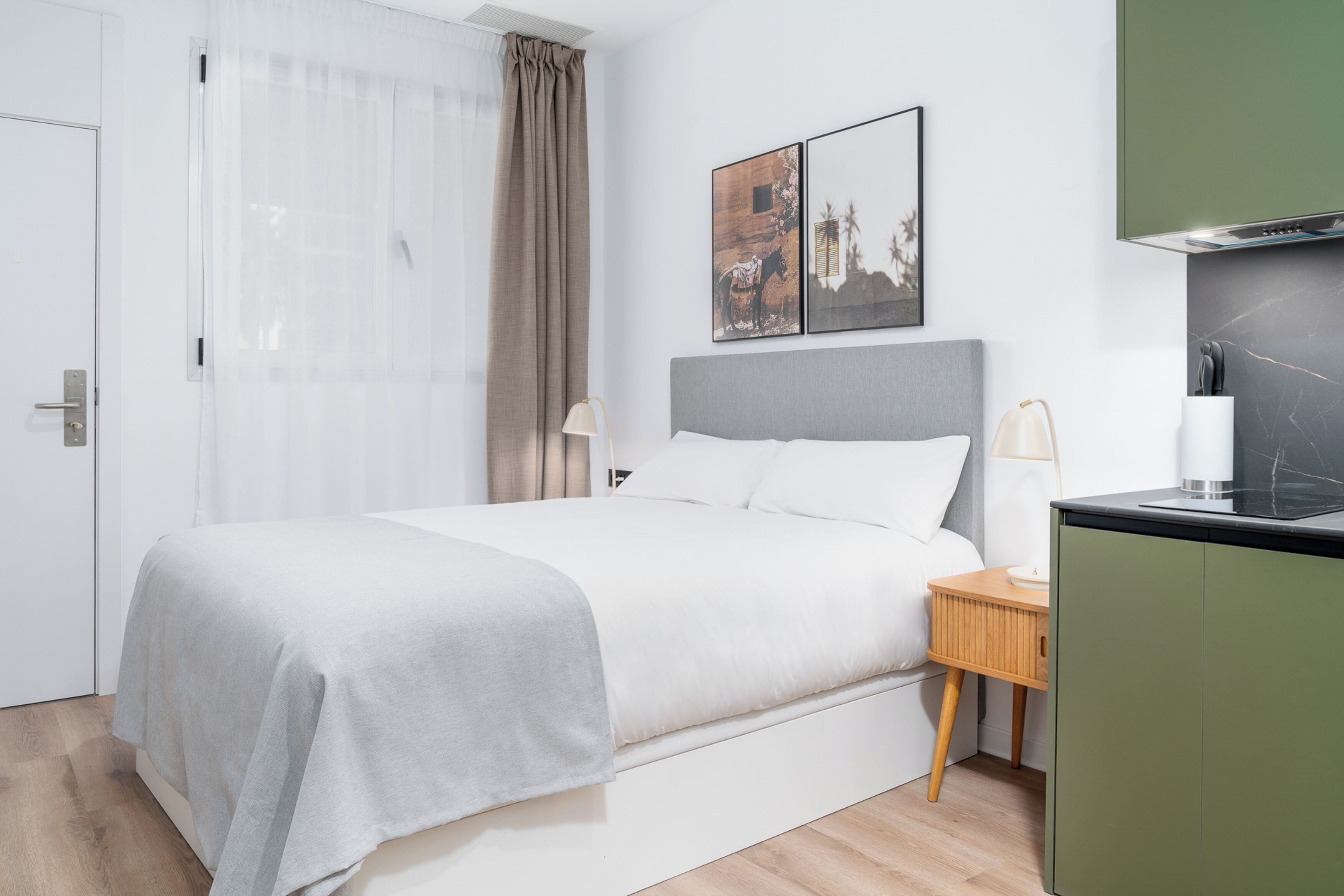 A modern hotel room with a bed, a nightstand, a lamp, a painting, and a kitchen area.