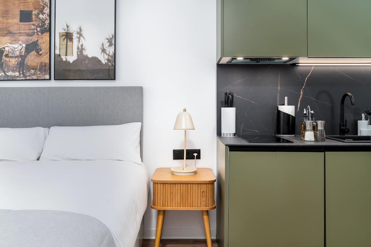 A modern bedroom with a bed, bedside table with a lamp, and a kitchen area with a sink and cabinets.