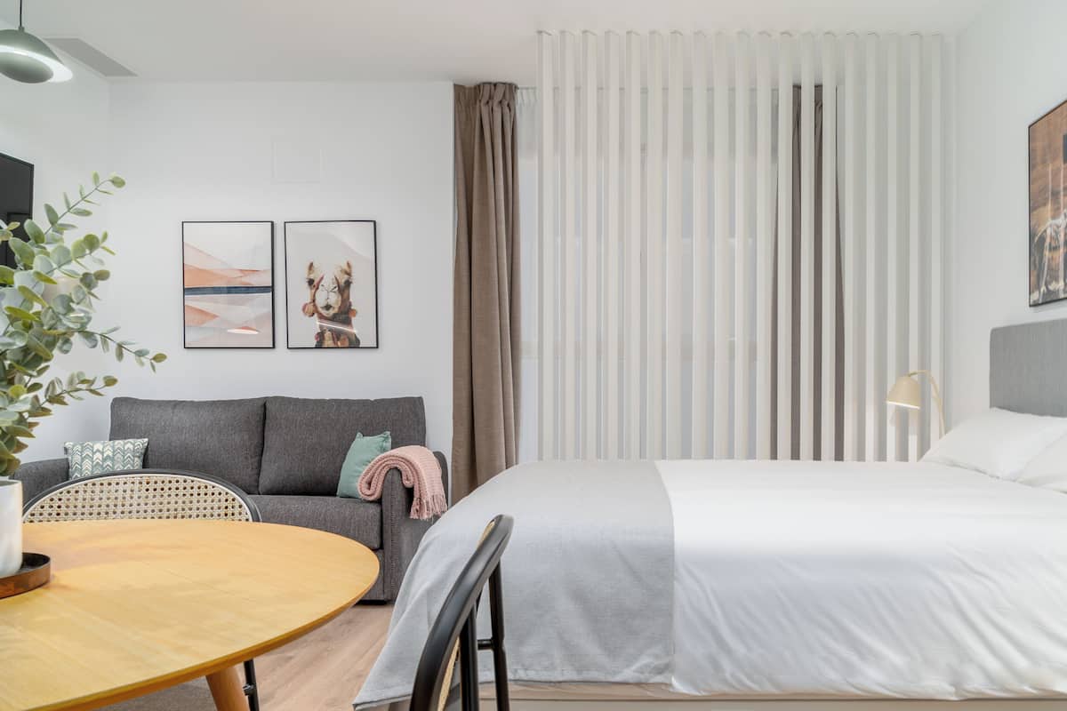 A modern hotel room with a bed, a sofa, a dining table, and a window with curtains.