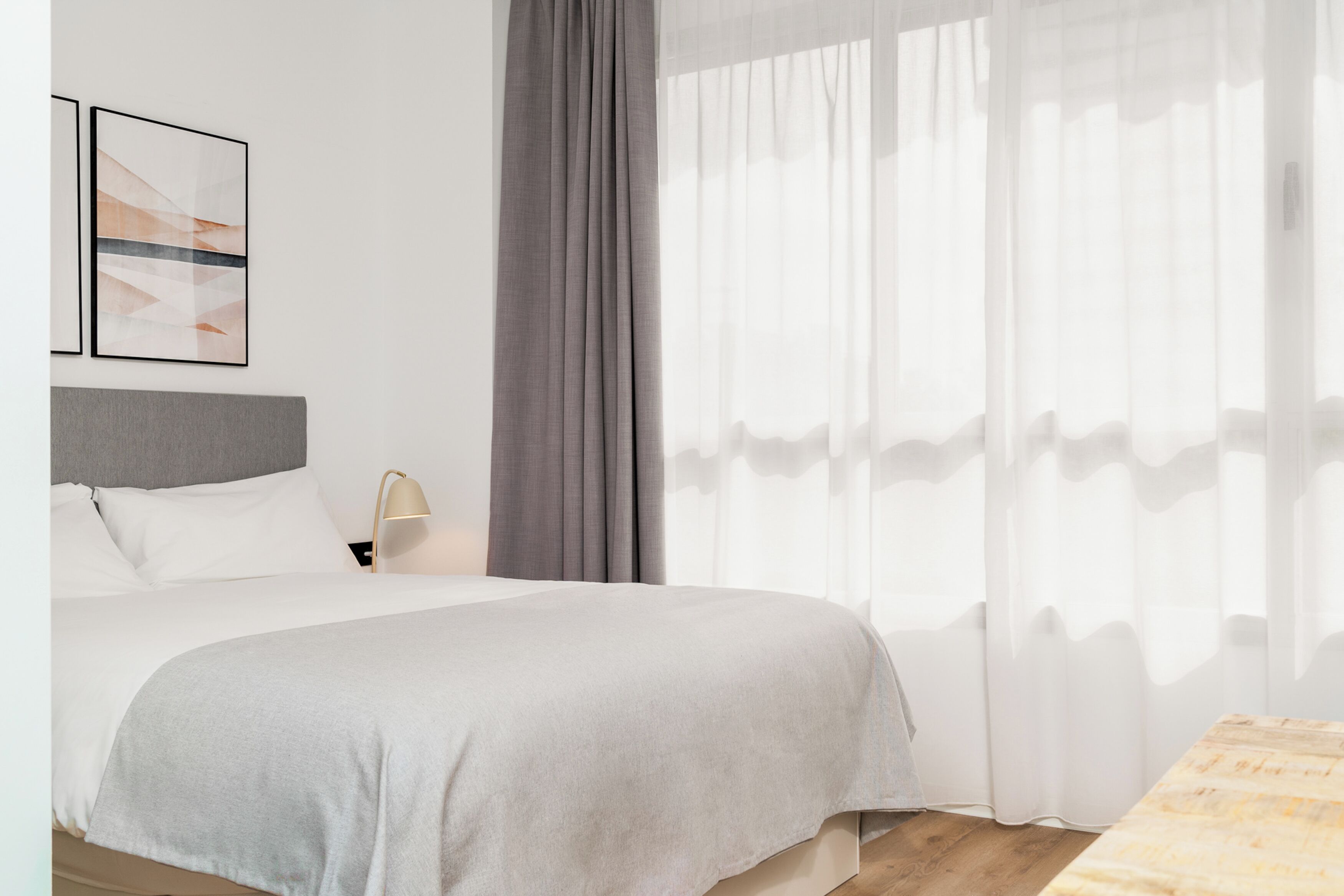 A hotel room with a bed, a bedside lamp, a window with sheer curtains, and a framed abstract artwork on the wall.