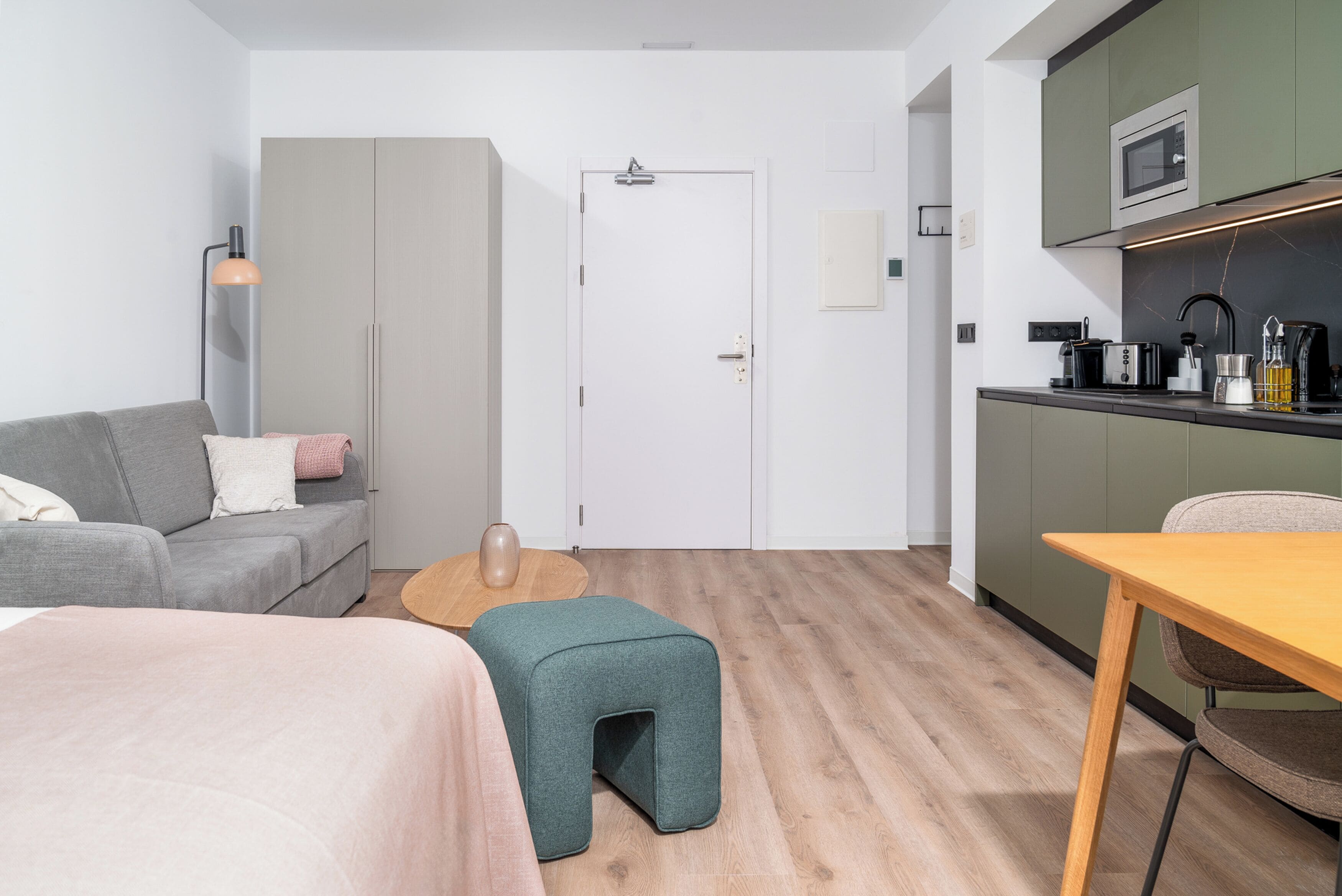 A modern, compact room with a grey sofa, a small green ottoman, a wooden coffee table, a wooden dining table, and a kitchenette with a microwave and toaster.