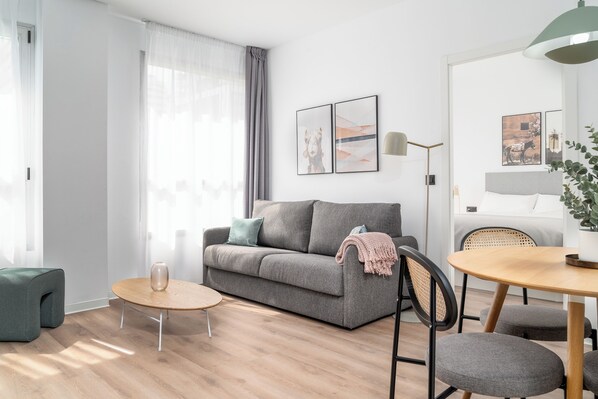 Uma sala de estar moderna com um sofá cinzento, uma mesa redonda de madeira e um banquinho verde pequeno.