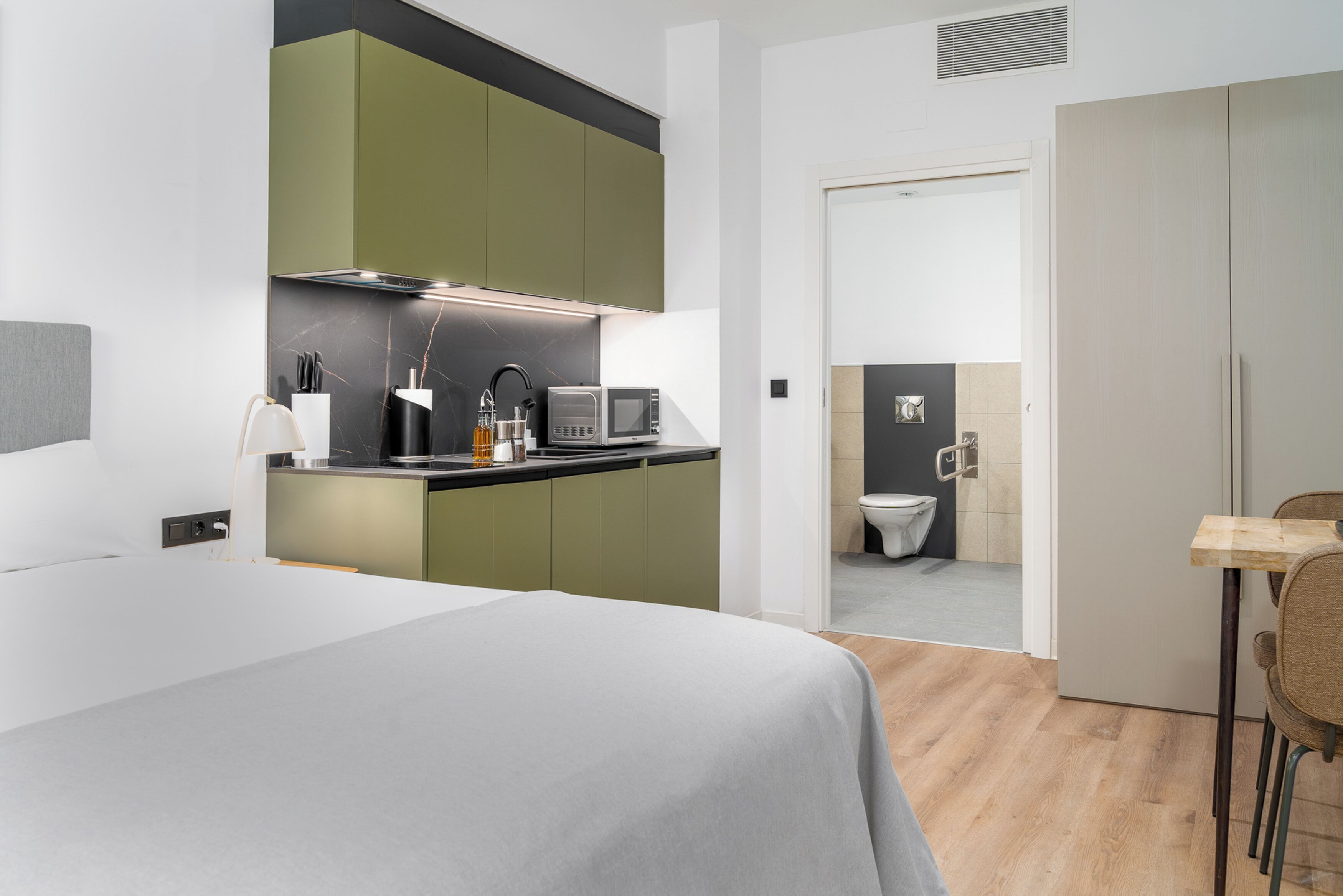 Een moderne slaapkamer met een groot bed, een kitchenette met groene kastjes, een magnetron en een kleine tafel met stoelen.