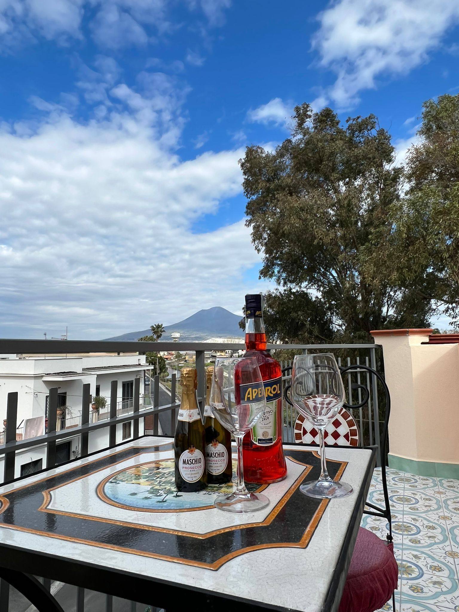 A table with bottles and glasses on a balcony overlooking a cityscape with buildings and a mountain.