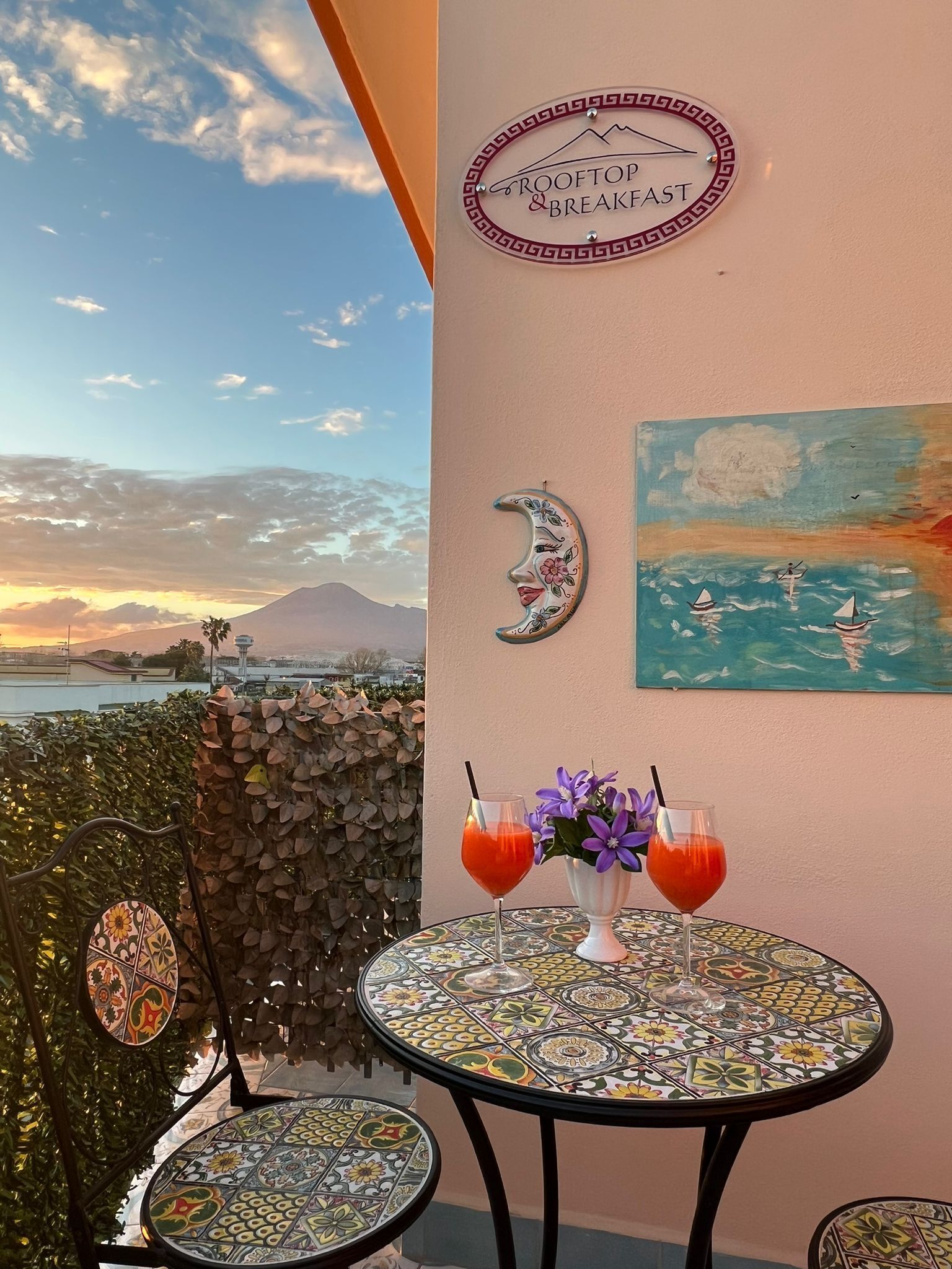 Varanda com uma mesa posta para dois, uma placa com a inscrição "Rooftop Breakfast" e uma pintura de veleiros.