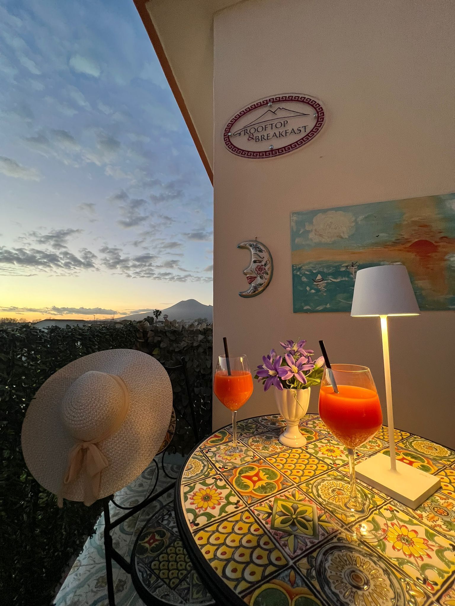 A balcony with a view of the sky and mountains, a table with drinks and a hat, and a sign that reads "ROOFTOP & BREAKFAST".