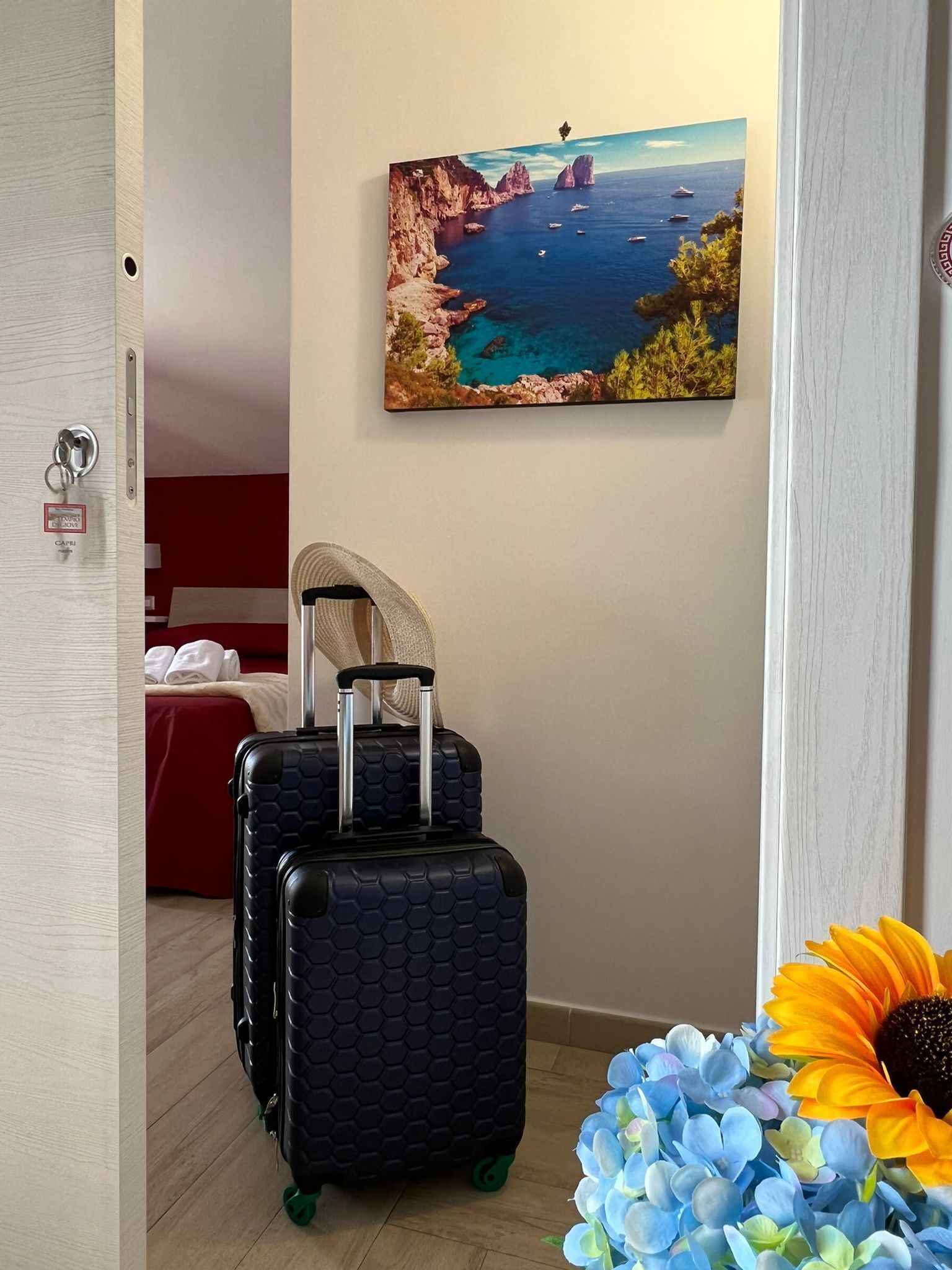 Una habitación con una cama, un cuadro de una escena costera, dos maletas y un adorno con girasol.