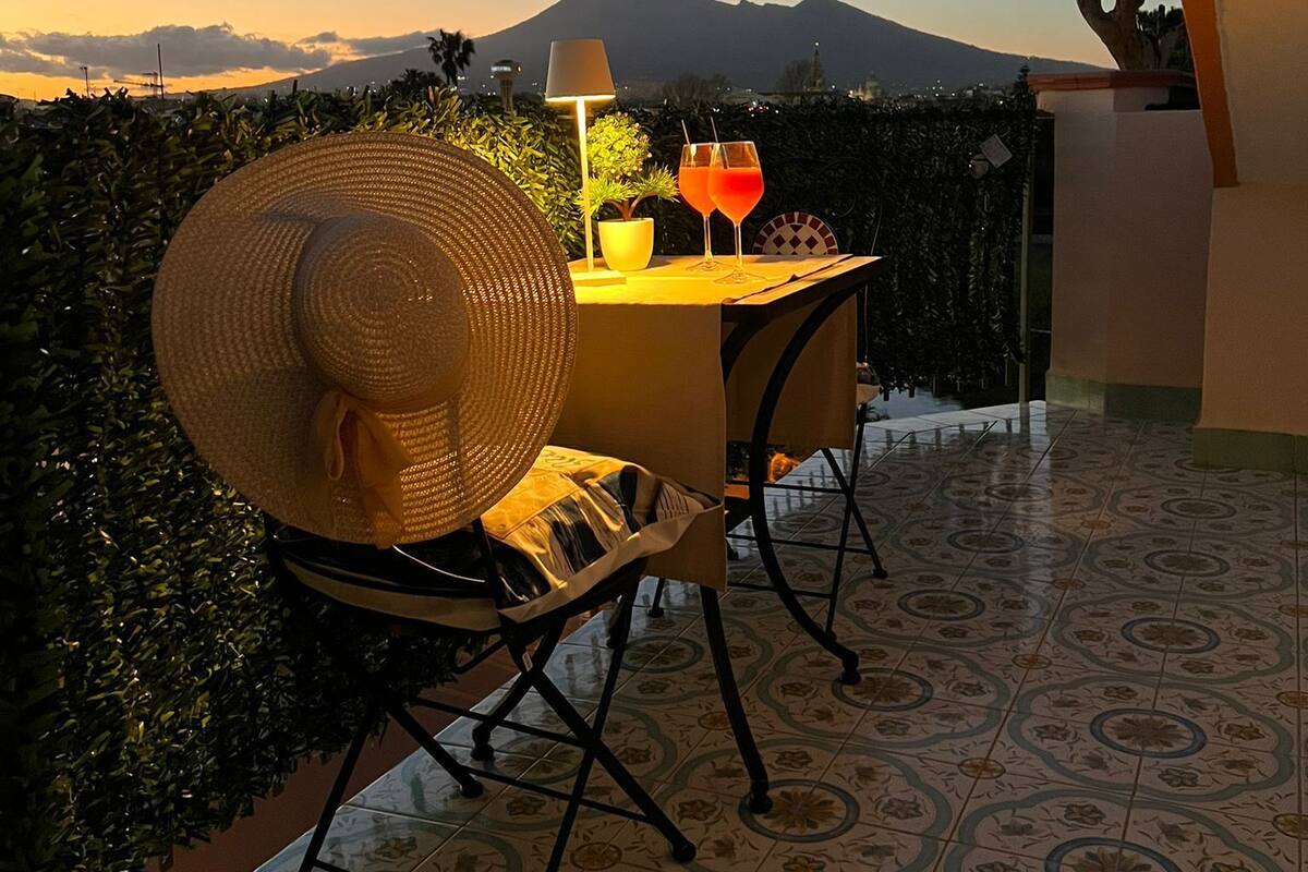 Un balcon avec une table dressée pour deux personnes, un grand chapeau en fibres tressées et deux boissons. On aperçoit une montagne au loin.