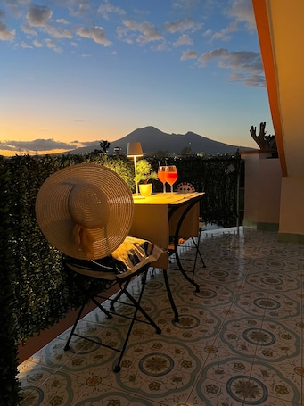 A balcony with a table set for two, a large woven hat, and two drinks. A mountain is visible in the background.
