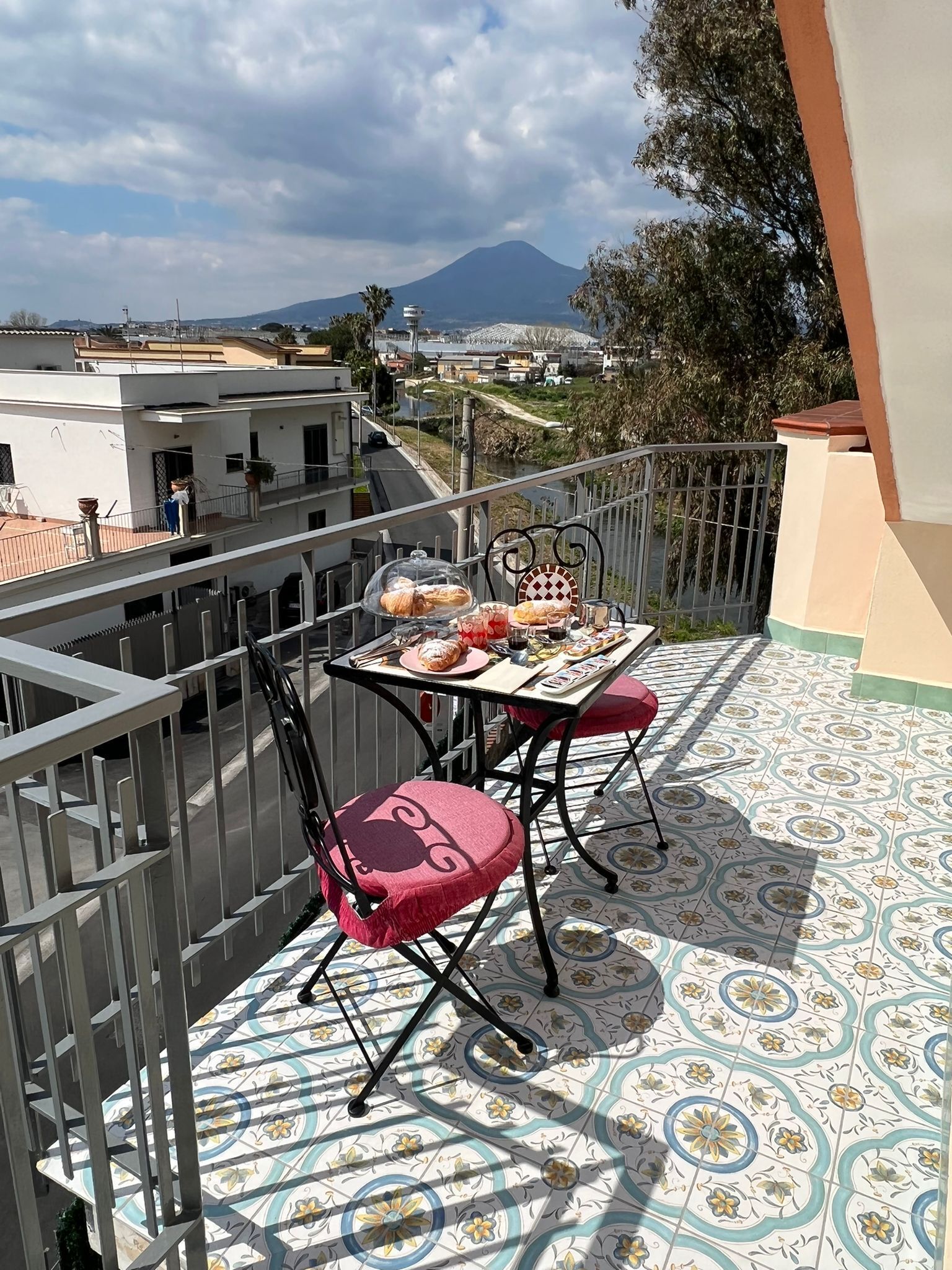 A balcony with a table set for two, featuring bread and pastries, and a chair with a cushion.
