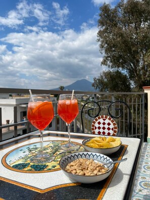 Ein Tisch mit zwei Gläsern roter Flüssigkeit, einer Schüssel Erdnüssen und einer Schale Chips auf einem Balkon mit Blick auf die Berge.