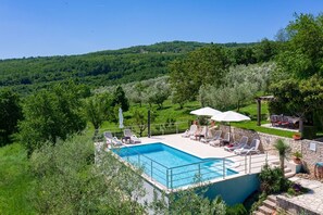 Une piscine rectangulaire bordée de chaises longues et d’ombrelles, entourée d’une végétation luxuriante et de collines.