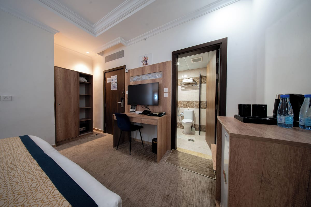 A hotel room with a bed, a desk with a chair, a television, a bathroom with a toilet and sink, and a minibar.
