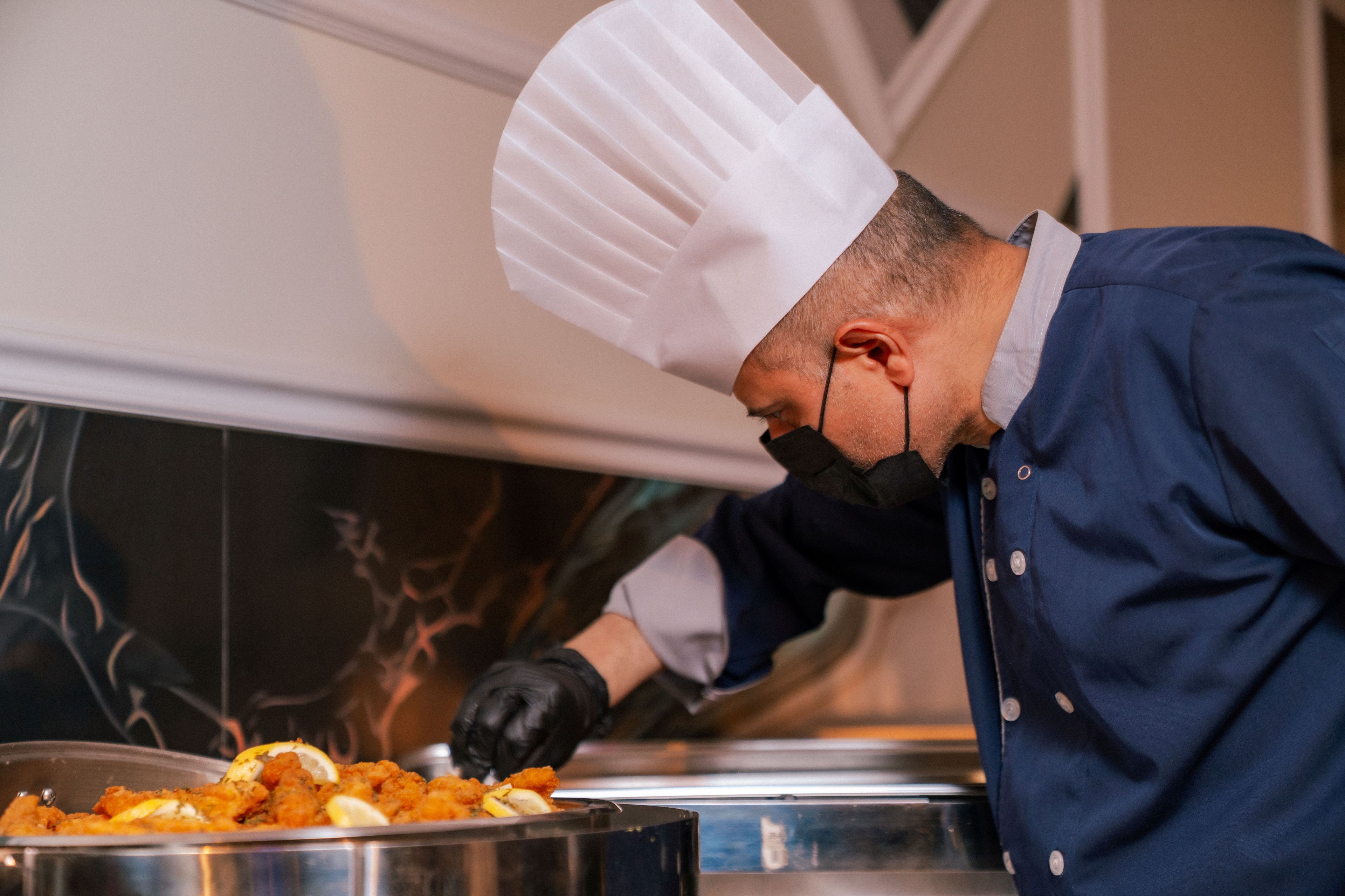 Um cozinheiro de uniforme e chapéu prepara alimentos em uma cozinha.