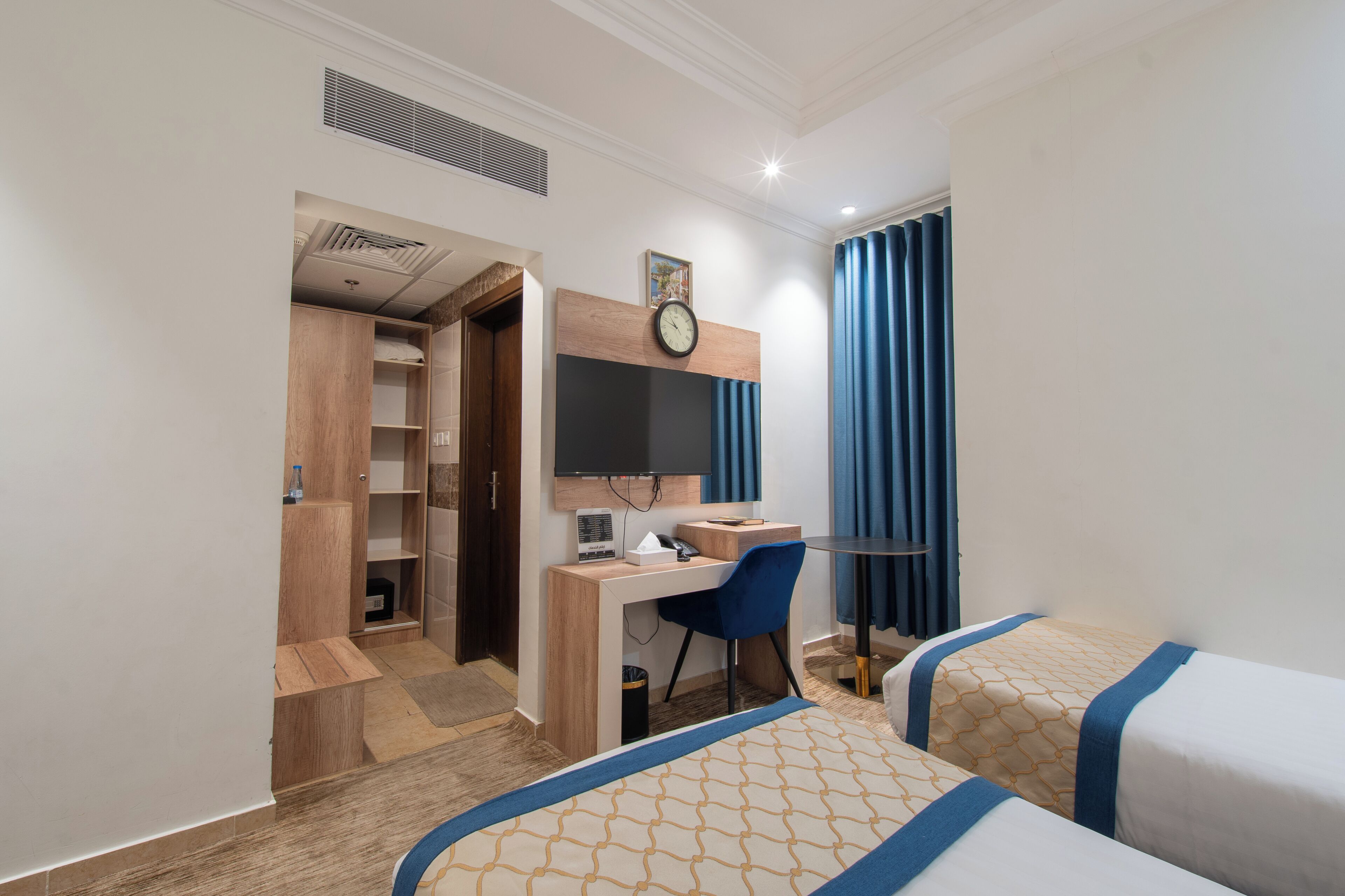 A hotel room with a desk, chair, two beds, a clock, and a wardrobe.