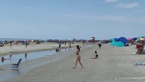 Una playa concurrida donde gente camina, se sienta y toma el sol.