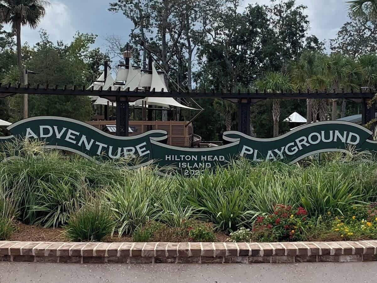 A sign for Hilton Head Island Playground with a wooden structure in the background.