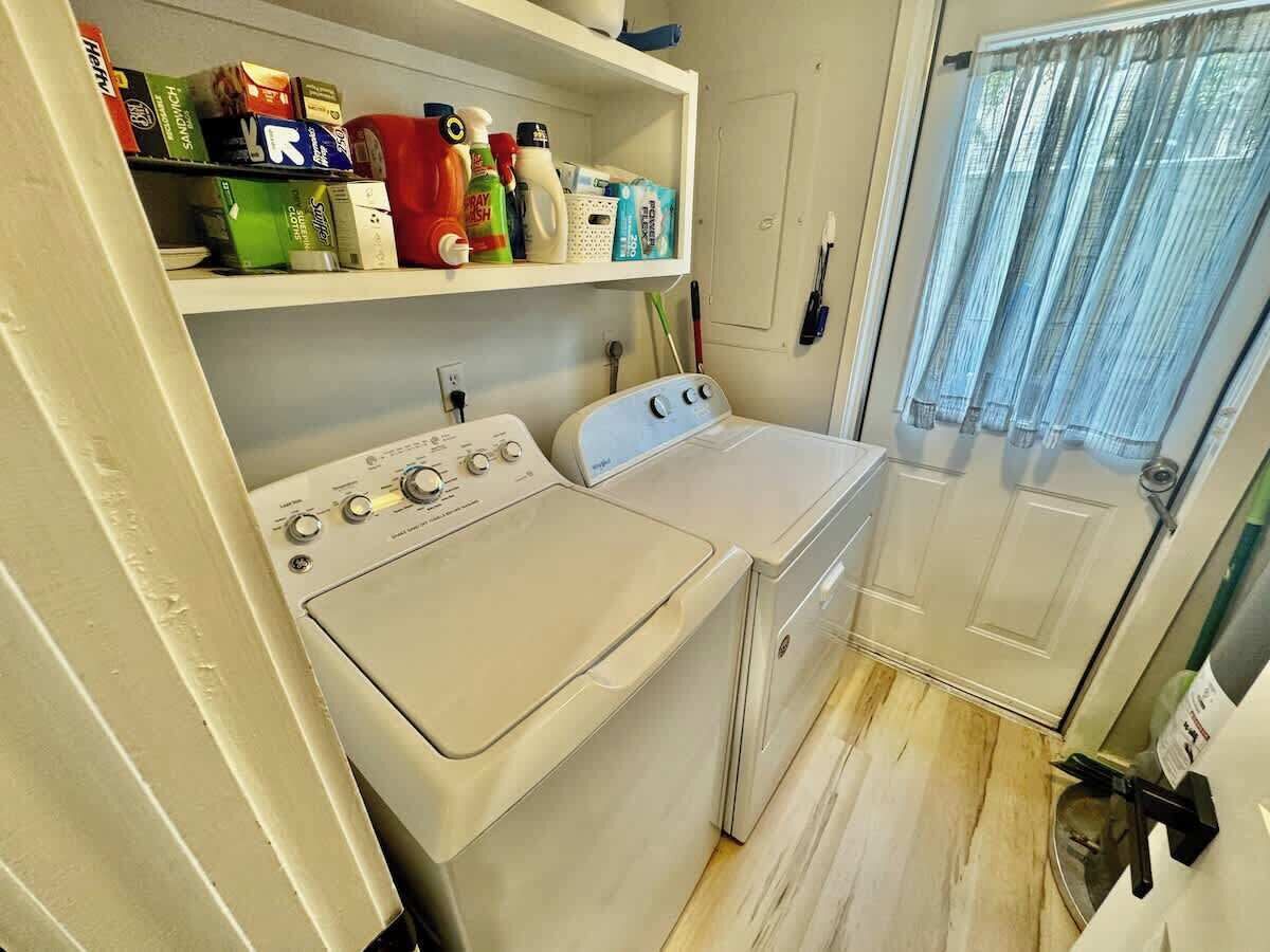 Un espace réduit dédié à la buanderie, comprenant un lave-linge et un sèche-linge, une étagère avec des produits de nettoyage et une porte munie d’un rideau.