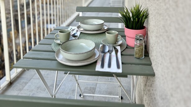 A balcony with a table set for dining, a potted plant, and a view of buildings in the background. - Spacious 2 Bedrooms Apartment With Acropolis View In Center of Athens (Athina)