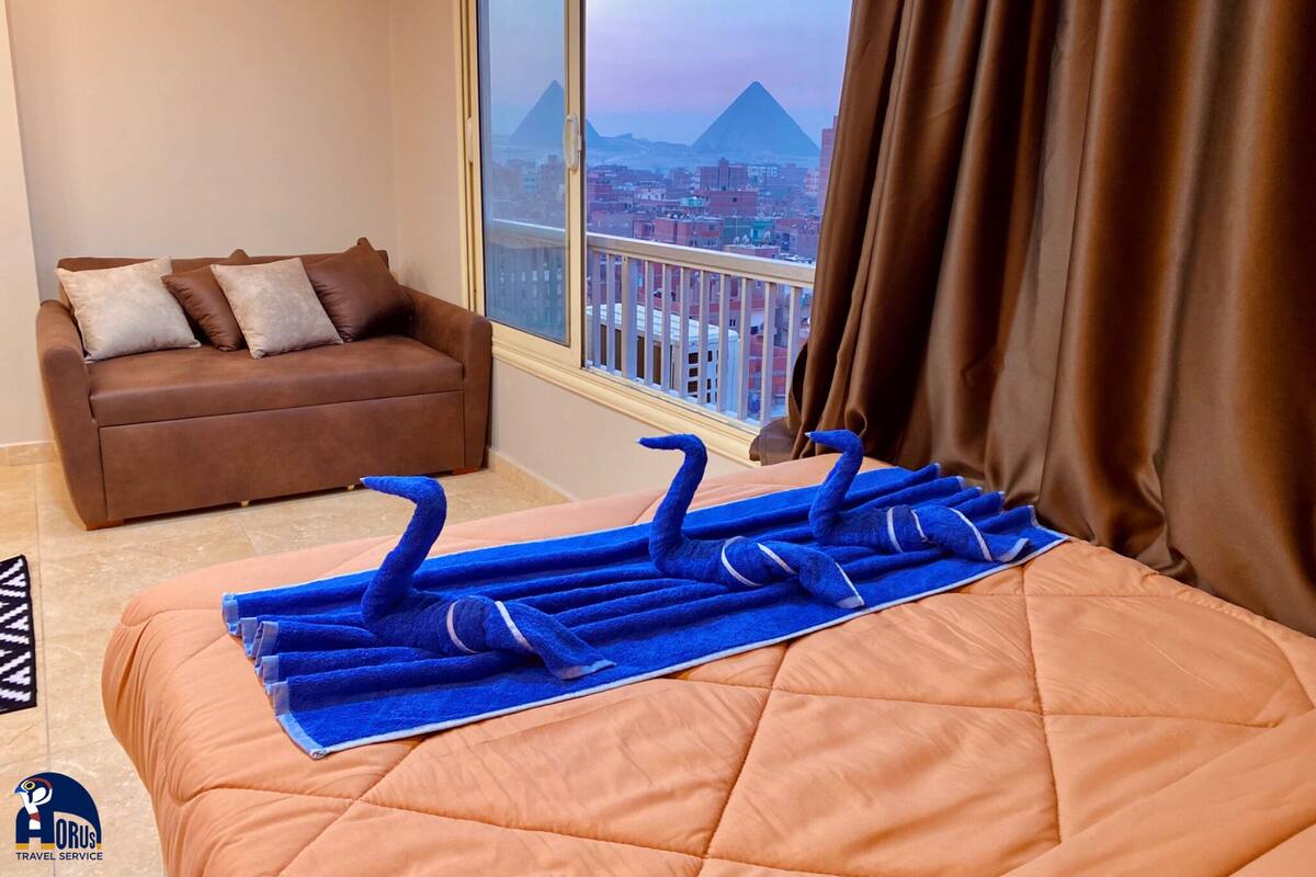 A hotel room with a bed, a brown sofa, a window with a view of a pyramid, and a balcony railing.
