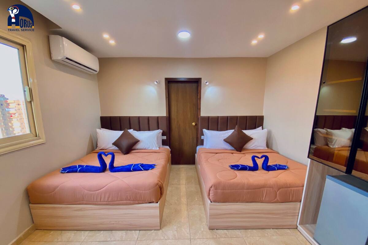 A hotel room with two beds, each with a swan-shaped towel, beige walls, and a window with a city view.