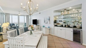 A modern kitchen with white cabinetry, a dining area with a white table and chairs, a chandelier, and a large window overlooking the ocean. - Pearl of Navarre 402 | Gulf front condo | Resort Amenities and More (Navarre Beach)