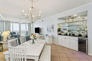 A modern kitchen with white cabinetry, a dining area with a white table and chairs, a chandelier, and a large window overlooking the ocean.