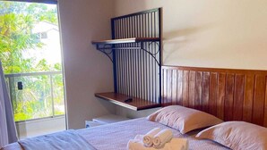 A bedroom with a bed, pillows, a wooden headboard, a window with a view of greenery, and a balcony. - Guarapari Park (Guarapari)