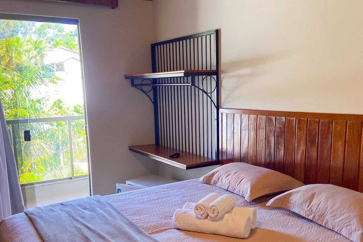 Une chambre à coucher avec un lit, des oreillers, une tête de lit en bois, une fenêtre donnant sur un paysage verdoyant et un balcon.