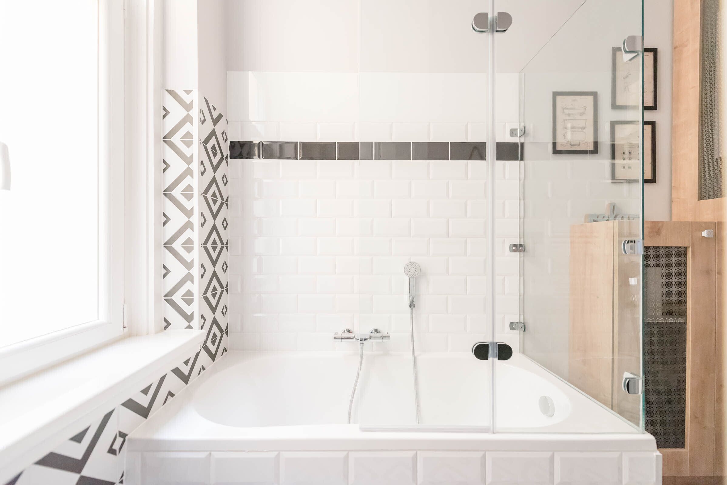Een moderne badkamer met een wit bad, een douche met glazen wanden, en een betegelde achterwand met een patroon.