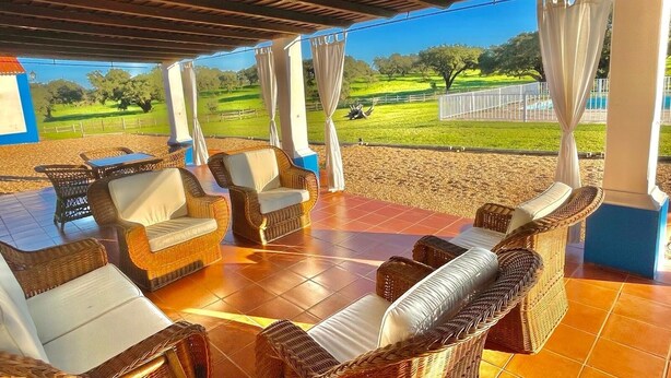 A covered outdoor seating area with wicker furniture and curtains, overlooking a green landscape.