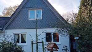 Una casa con un techo pronunciado, un ventanal pequeño con marco blanco y un comedero para pájaros colgado de un poste de madera.