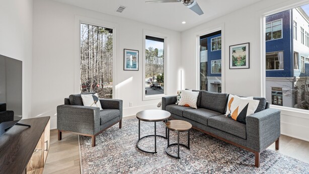 A modern living room with a grey sofa, two armchairs, a coffee table, and a TV. Large windows offer views of trees and buildings. - Plutus' Playhouse (Palmetto)